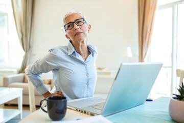 Portrait of senior stressed woman sitting at home office desk in front of laptop, touching aching...