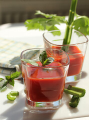 Cocktails. Bloody Mary. Red drink made from tomatoes, lemon juice, vodka, celery, spices in a clear glass on a white table. Rustic. Tomatoes, basil, sunlight, vertical