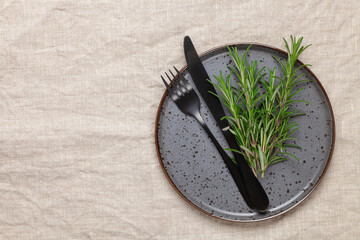 Fototapeta na wymiar Black plate, cutlery and napkin with a sprig of rosemary on textile table. Top view. Table setting.