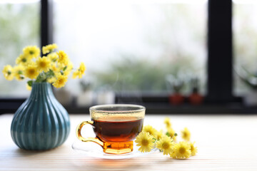 Small Chrysanthemum flower tea glass cup and flower pot on wooden table in front of glass window
