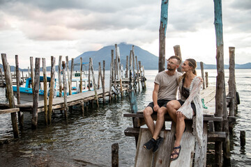 travel couple sitting on old dock on lake atitlan guatemala with volcano in background