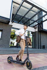 full length of businessman in formal wear with leather bag standing near e-scooter and building