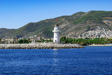 Fototapeta na wymiar Alanya lighthouse with the Turkish flag on the roof against the background of a mountain panorama of the Mediterranean coast. Seascape with tropical resort town among the hills on the horizon