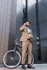 Fototapeta na wymiar low angle view of businessman talking on smartphone and holding paper cup near bicycle and building