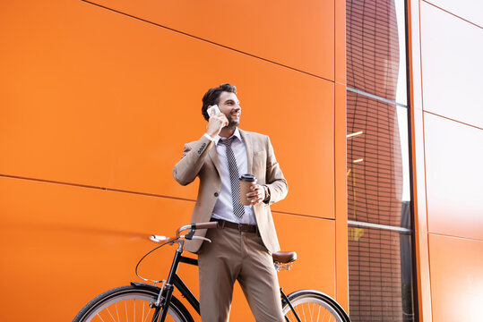 cheerful businessman talking on smartphone and holding paper cup near bicycle and building with orange walls