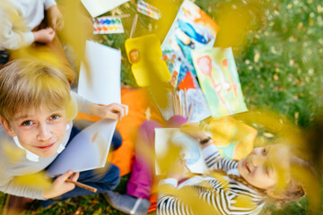 Cute child with funny face looking up at camera. Children, boy and girlsitting on green grass in creative chaos palette watercolor paints, brush, drawings. Painting outdoor. Top view. Love friendship.