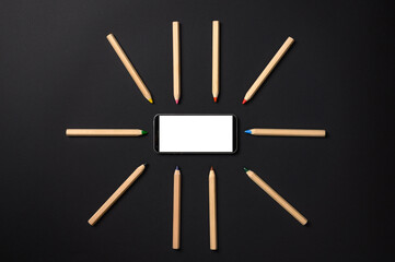 Pencils lie around the smartphone on a black background. Smartphone with blank white display