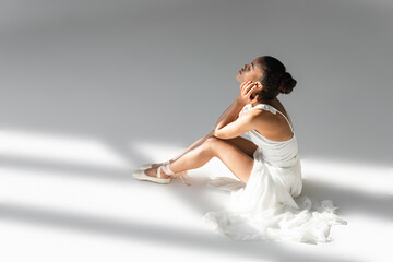 Fototapeta na wymiar graceful african american ballerina in dress sitting on floor on white background