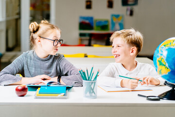 Smiling schoolgirl talking with friend, looks at each other, smiling while writing in notebook in classroom at desk