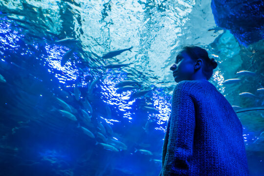 Underwater life, tourism, education and entertainment concept. Portrait of woman looking at fish vortex in large public aquarium tank at Oceanarium with blue low light illumination - low angle view