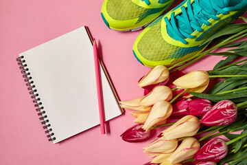 Spring sport composition with running shoes, red and yellow tulips and open blank notebook on pink background, copy space. Sports concept. Top view, flat lay