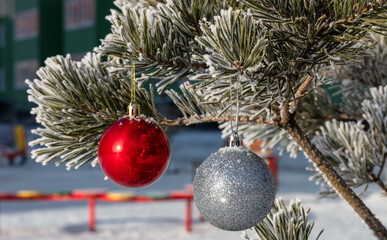 Close up shot of a glittering white and blurred shiny red Christmas balls hanging off a Christmas fir tree outside, all partially covered in snow. Blurred building as a backdrop
