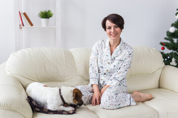 Happy young woman wearing pajama with lovely dog in living room with christmas tree. Holidays concept.