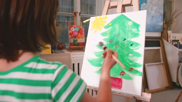 A child paints a Xmas tree decorations on the canvas. Christmas and Advent art lessons for kids focus on multiple art elements and principles, and exemplify the spirit of the season. No face, indoor.