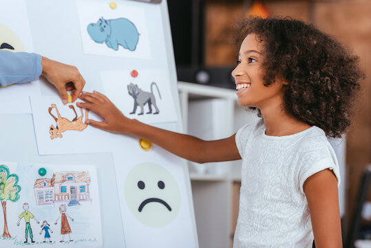 Cheerful african american girl touching picture on whiteboard near hand of psychologist on blurred background