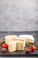 Cheddar cheese and various types of cheese with rosemary and tomatoes on black slate board on a black concrete background. Side view, copy space