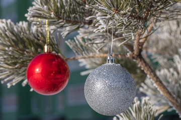 Close up shot of a glittering white and blurred shiny red Christmas balls hanging off a Christmas fir tree outside, all partially covered in snow. Blurred building as a backdrop