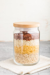 glass jar with different kinds of rice poured in layers on a gray and white background. side view, close up.
