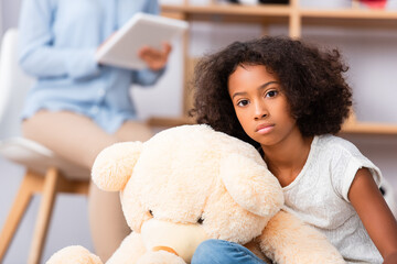 Lonely african american girl looking at camera near teddy bear with blurred psychologist on...