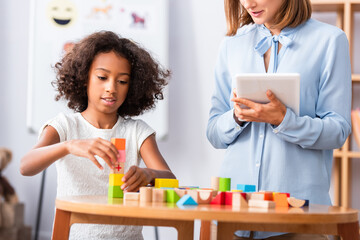 African american girl playing with wooden blocks near psychologist holding digital tablet with...