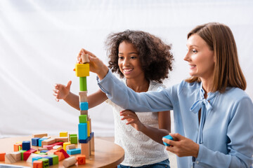 Happy psychologist building tower with wooden blocks near cheerful african american girl at coffee table