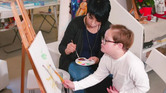 A boy with Down Syndrome sits by a canvas and an easel, a supporting student assistant nearby. Multisensory activities during art lesson and materials work well to adaptation. 
