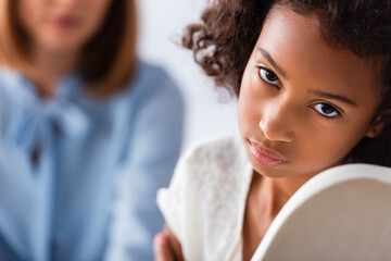Depressed african american girl with autism looking at camera during consultation with blurred psychologist on background