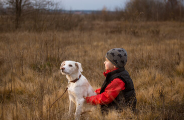 toddler boy walks in nature with his white young retriever dog, sit in an embrace among dry grass. The joy of communicating with a four-legged friend, a beloved pet