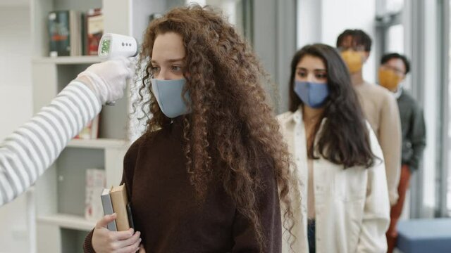 Diverse students wearing masks standing in line at entrance, getting their temperatures checked by unrecognizable person with contactless thermometer, Caucasian girl passes, Mixed-Race one not