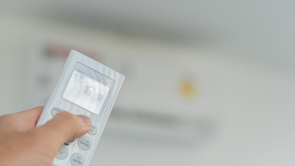 The hand on the remote control is about to drop the too cold air conditioner in the room with blurry background.