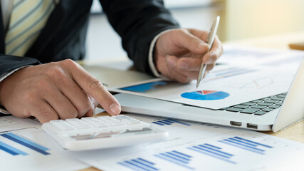 Hand pointing at the calculator, beside, holding the pen, pointing at the graph on the businessman's laptop on the table with graph and orange light.