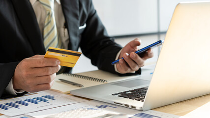 Online payment man's hand holds credit card and uses a smartphone to shop online and on the table a laptop with a graph