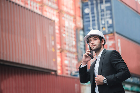 Handsome business man wears white hardhat and holding cellphone or smartphone with stacked cargo container. Logistic warehouse business and transportation concept.