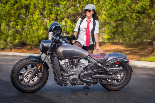 Woman next to a motorcycle dressed in vintage clothing including a red tie and cap