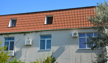 A small residential house with a mansard red clay roof and attic or mansard windows, air conditioner outdoor units on a stucco gray wall.