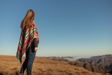 a girl in a colored national poncho stands and looks into the distance. girl on the background of the Caucasian mountains in full growth