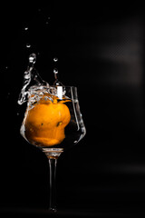 AN ITALIAN  TANGERINE FALLS AND SPLASHES INTO A WINE GLASS FLOOR OF WATER CREATING SPLASHES AND PLAYS OF LIGHT ON A BLACK ORANGE BACKGROUND