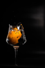 AN ITALIAN  TANGERINE FALLS AND SPLASHES INTO A WINE GLASS FLOOR OF WATER CREATING SPLASHES AND PLAYS OF LIGHT ON A BLACK ORANGE BACKGROUND