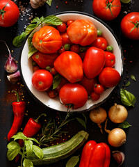 Various types of tomatoes in a bowl and fresh vegetables and herbs around on a dark background, top...