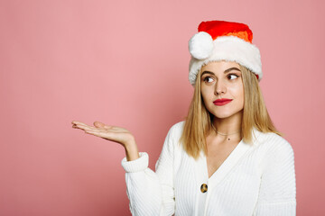 Smiling young 20s Santa woman in sweater, Santa hat pointing with palm to the side on mock up copy space isolated on pink background, studio portrait. Happy new year celebration merry holiday concept