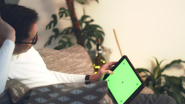 Close up of man lying down on sofa watching digital tablet with chroma key during winter eve. Green screen display. Digital Concept for remote video call, video on demand, websites and application.