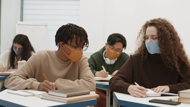 Medium close-up of African male student in glasses writing off lecture of female Caucasian groupmate, girl moving notebook away from him. People wearing masks, sitting by desks in classroom