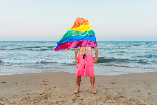 Man standing on the beach with gay pride flag covefring his face