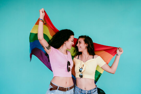 Happy lesbian couple holding rainbow flag looking at each other while standing against wall