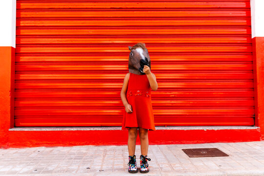 Little girl with a horse's head and a red dress standing in front of red roller shutter