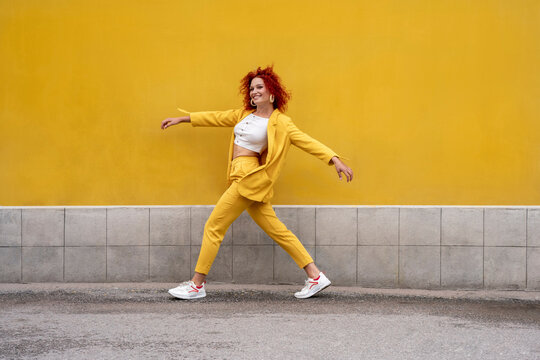 Energetic young woman in yellow suit running and jumping in front of yellow wall