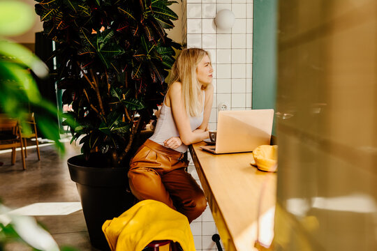 Attractive European Woman Is Sitting Alone At A Table In A Cafe. She Seems To Be Looking At Her Laptop Doing Work And Thinking. It Is Indoors With A Bright Background With Plants.