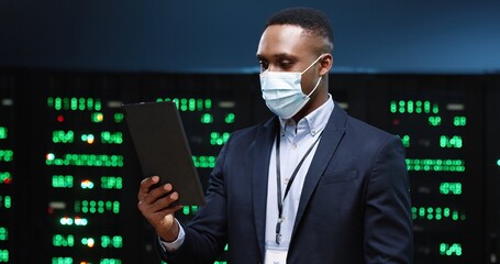 Close up portrait of happy African American man in medical mask standing among servers videochatting on tablet while checking big data processors. Male analytic speaking in video in data storage