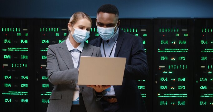 Portrait of Caucasian female system administrator in mask typing on laptop and speaking with African American male security engineer standing in datacenter. Big data protection and digital information - Powered by Adobe