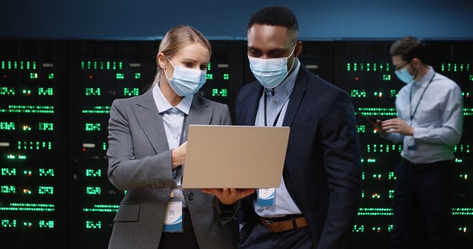 Portrait of Caucasian female system administrator in mask typing on laptop and speaking with African American male security engineer standing in datacenter. Big data protection and digital information